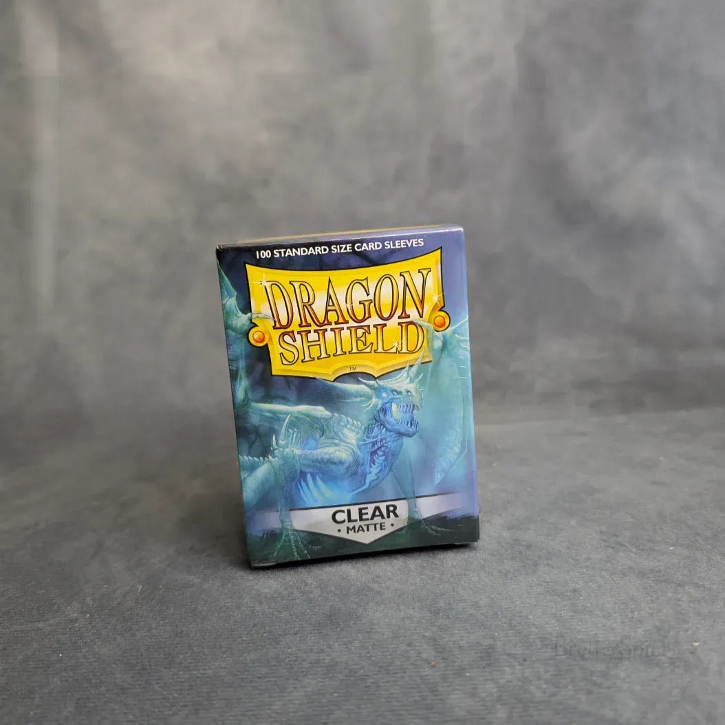Eine Schachtel von Brett&Spiel Dragon Shield: Matte - Clear (100) Kartenhüllen Standard sitzt auf einer grauen Oberfläche, zeigt Fantasy Art mit einem blauen Drachen und einem gelben Logo und bietet optimalen Schutz für Sammelkarten.