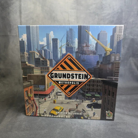 Das Bild zeigt das Schachtelcover von Grundstein von Metropolis von Kobold Spiele, das den Bau von Städten mit Kränen und Wolkenkratzern zeigt, was das Polyomino-Bauthema des Spiels widerspiegelt.