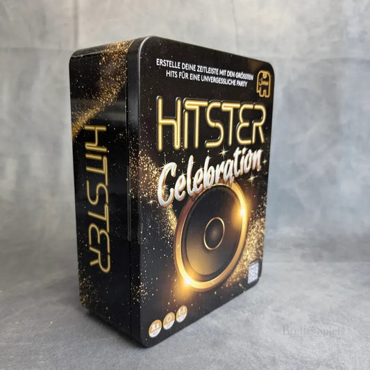 Eine schwarz-goldene Schachtel mit der Aufschrift "Hitster: Celebration" von Jumbo Spiele ist mit einem Lautsprecher, Musiknoten und einem deutschen Text versehen, der es als Musikspiel für den Partyspaß ausweist. Die aufrecht stehende Box steht auf einer neutralen Fläche.