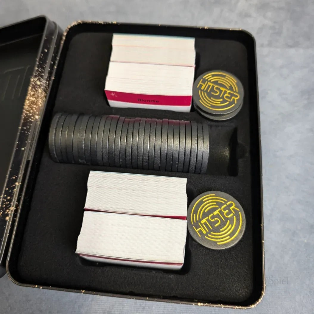 Hitster: Celebration von Jumbo Spiele kommt in einer Metalldose mit gestapelten schwarzen Pokerchips, zwei gelb-schwarzen HINTSTER-Spielsteinen und vier Stapeln rot-weißer Karten - ideal für deine nächste Party!.