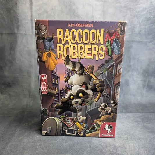 Die Raccoon Robbers-Box von Pegasus Spiele zeigt Cartoon-Waschbären, die Essen stehlen und über Wäscheleinen, Mülltonnen und Dächer fliehen. Der fette gelbe Titel und das verspielte Artwork machen das Spiel zu einem echten Hingucker.
