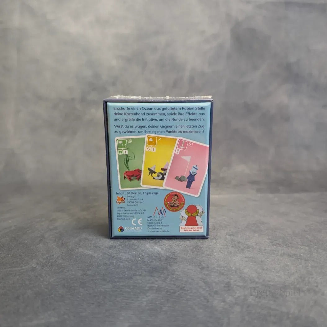 Eine kleine Meer, Salz & Papier + Extra Salz Erweiterung Box von MM-Spiele steht aufrecht auf einer neutralen grauen Oberfläche. Auf der Rückseite befinden sich drei illustrierte Karten mit Tieren und Zahlen sowie ein deutscher Text zu diesem Karten-Sammel-Spiel.
