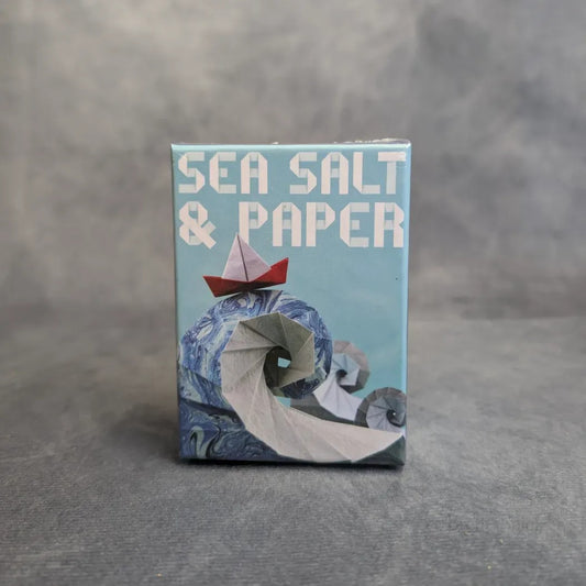 Eine Schachtel von Sea, Salt & Paper + Extra Salt Erweiterung von MM-Spiele zeigt ein rot-weißes Origami-Boot auf einer blau-weißen Welle vor einem hellblauen Hintergrund, der den einzigartigen Stil dieses Kartenspiels einfängt.