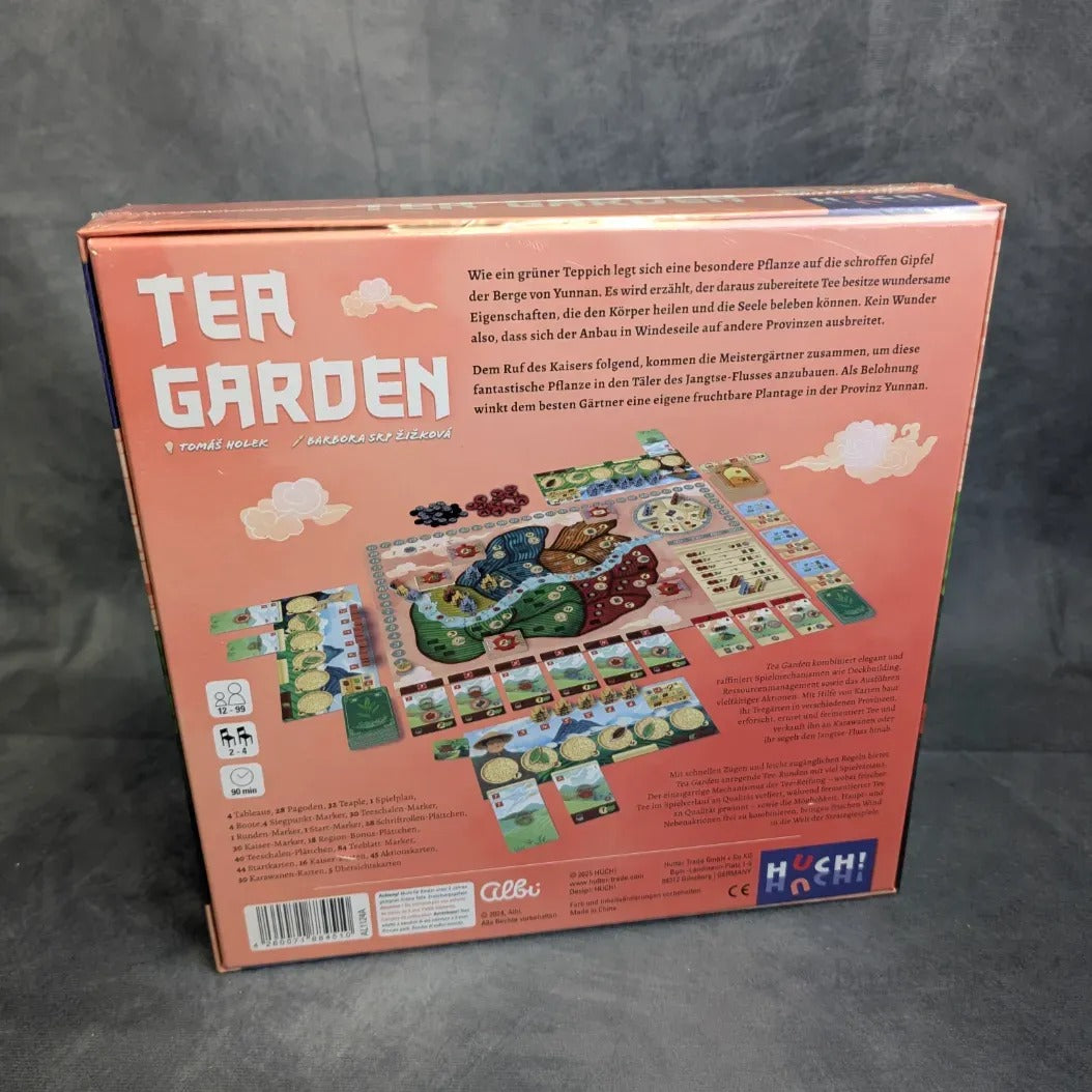 Die Rückseite der Schachtel des Brettspiels Tea Garden aus dem Huch Verlag zeigt einen lebendigen Aufbau mit Karten, Spielsteinen, Plättchen und Spielbrett, dazu deutsche Texte über den Teeanbau und Logos, alles vor einem rosa Hintergrund.