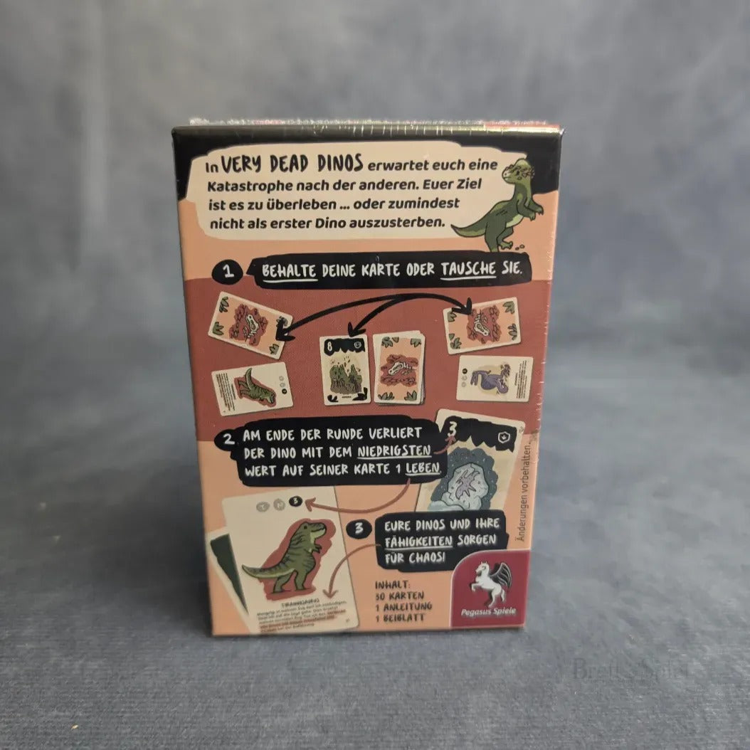Rückseite der Schachtel des Very Dead Dinos - TRex Kartenspiels von Pegasus Spiele, mit Cartoon-Dinosauriern und Karten auf einer grauen Oberfläche. Die deutsche Anleitung beschreibt die Schritte dieses lustigen Dinosaurier-Überlebensspiels.