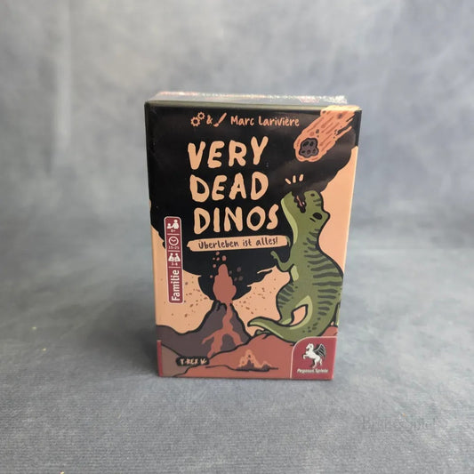 Das Kartenspiel The Very Dead Dinos - TRex von Pegasus Spiele zeigt verspielte Cartoon-Dinosaurier, einen ausbrechenden Vulkan und einen Meteor, mit kräftigen orangefarbenen, grünen und schwarzen Illustrationen und Texten in Deutsch und Englisch.