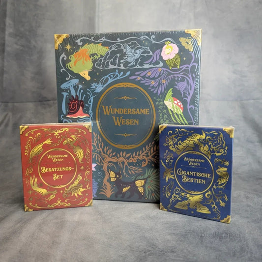 Drei Strohmann-Spiele-Schachteln mit Fantasy-Themen - "Wundersame Wesen" (groß), "Besatzungs-Set" (rot) und "Gigantische Bestien Erweiterung" (blau) - mit verschnörkelten Golddetails auf grauem Hintergrund.