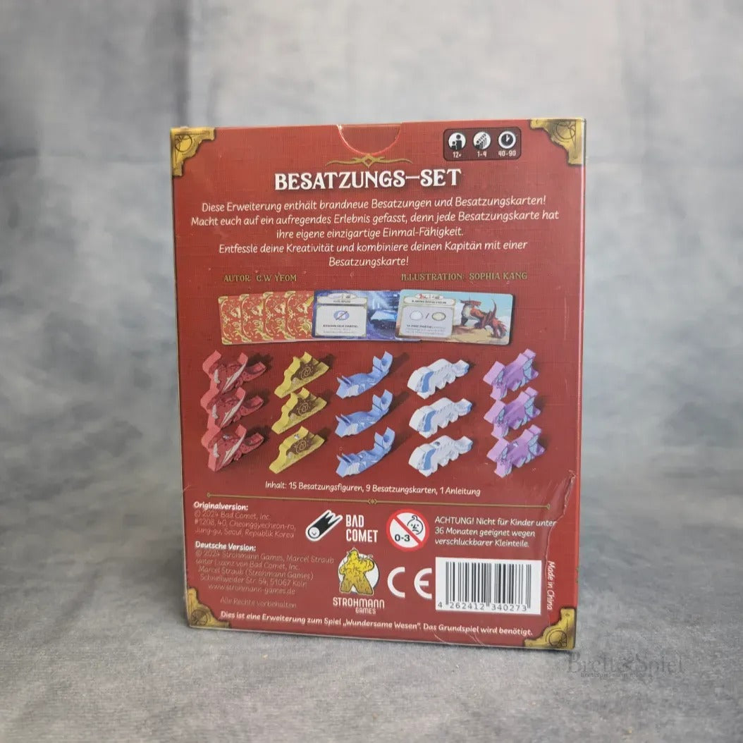 Die Rückseite des Wundersame Wesen + Besatzungs-Set + Gigantische Bestien Erweiterung von Strohmann Spiele zeigt Komponenten wie rote, violette und blaue Schiffsfiguren, Karten und Plättchen. Ein deutscher Text beschreibt das Set; Sicherheitshinweise erscheinen am unteren Rand.