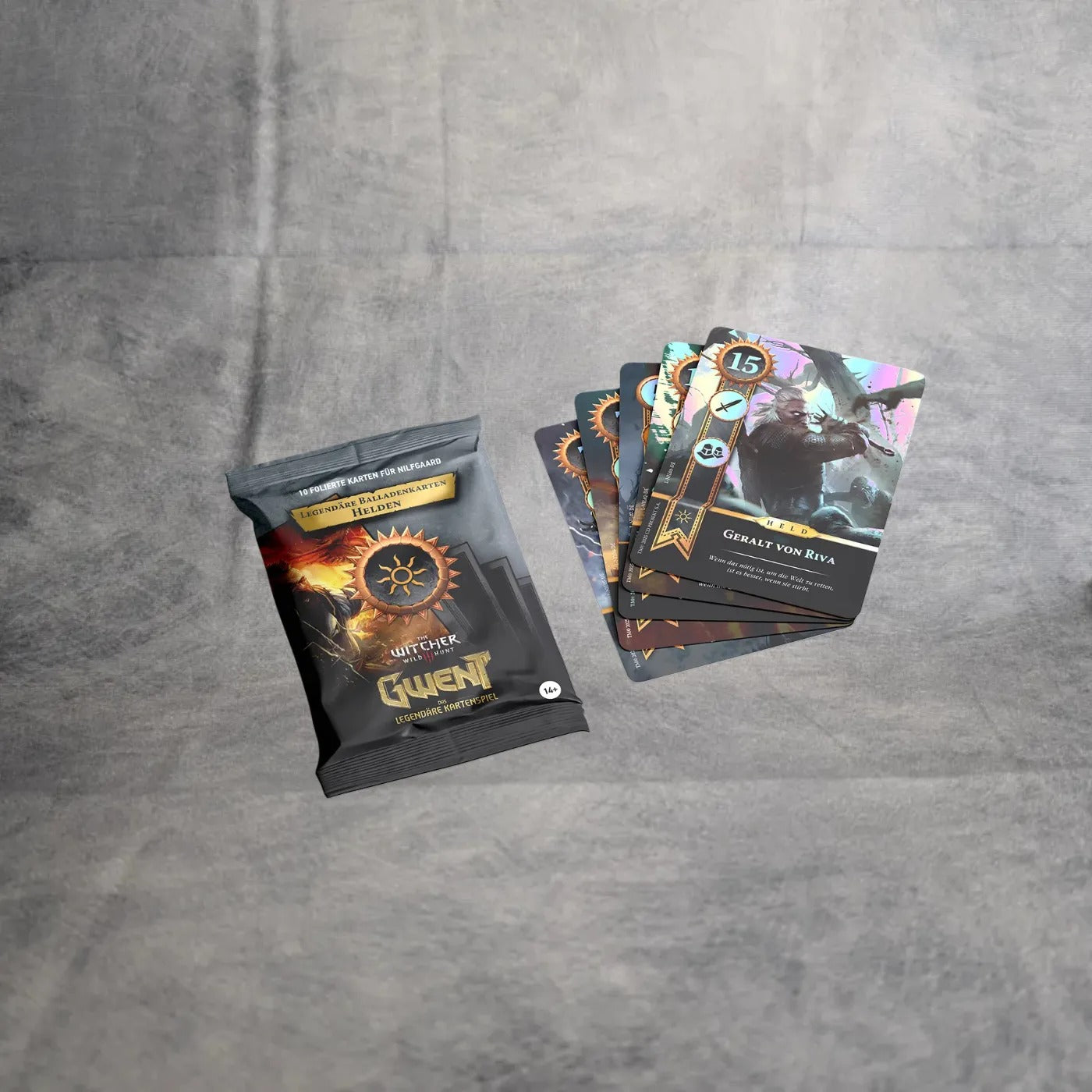 Ein Päckchen Gwent - Legendäre Balladenkarten von Asmodee liegt auf einer grauen Fläche neben mehreren aufgefächerten Karten, darunter eine mit dem Krieger-Artwork von Great Von Riva.