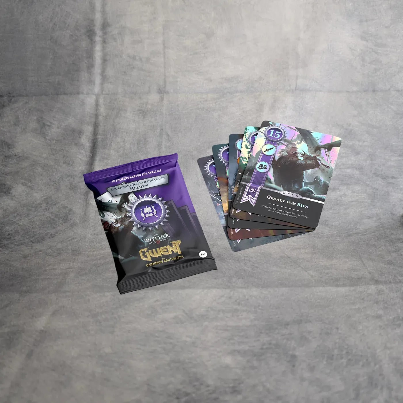 Eine versiegelte Asmodee Gwent - Legendäre Balladenkarten Packung ruht auf einer grauen Oberfläche neben fünf illustrierten Karten, wobei die oberste Karte Great Von Rivia aus dem The Witcher-Universum zeigt.