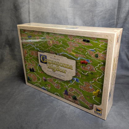 Ein Hans im Glück Carcassonne Big Box 3.1 Set steht aufrecht auf einer grauen Oberfläche und zeigt ein lebendiges Titelbild mit Landschaften, Straßen, Städten, Meeples und mehreren Erweiterungen.