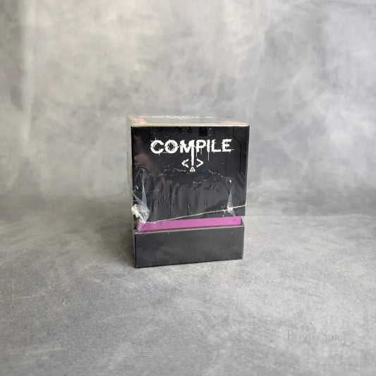 Eine schwarze Schachtel von Pegasus Spiele mit der Aufschrift "Compile" und einem Codesymbol auf der Vorderseite steht auf einer strukturierten grauen Fläche und ist teilweise von einer durchsichtigen, leicht zerknitterten Plastikfolie bedeckt.