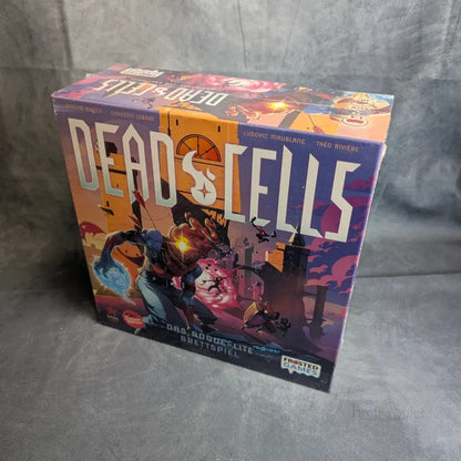 Eine Schachtel mit Dead Cells von Frosted Games ruht auf einer grauen Oberfläche. Das farbenfrohe Cover zeigt einen gepanzerten Helden mit einem leuchtenden Schwert vor einer stilisierten Stadt, die das schurkenhafte Dungeon-Crawling-Abenteuer des Spiels widerspiegelt.