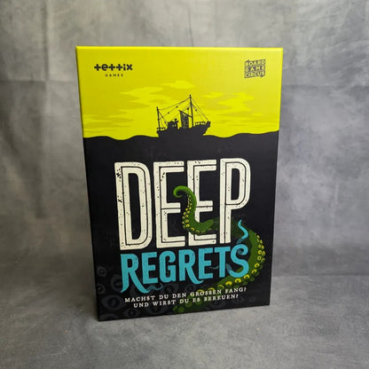 Die Schachtel des Brettspielzirkus für Deep Regrets zeigt ein Fischerboot, einen grünen Tentakel und einen gelb-schwarzen Hintergrund, der an düstere Folklore erinnert, mit dem Untertitel auf Deutsch.