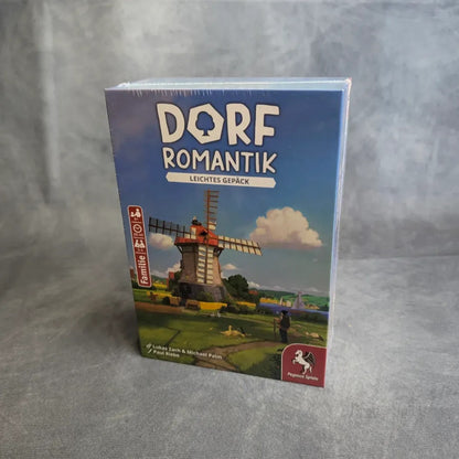 Ein verpacktes Brettspiel, Dorfromantik: Leichtes Gepäck von Pegasus Spiele, steht aufrecht auf einer grauen Fläche. Das Cover zeigt eine malerische Landschaft mit einer Windmühle - die kompakte Version des beliebten kooperativen "Legespiels".