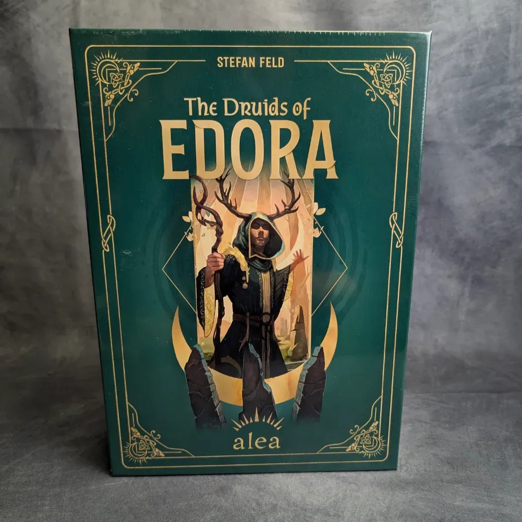 Das Cover der Box The Druids of Edora von Pegasus Spiele zeigt einen Druiden mit geweihtem Stab und Kapuzenmantel vor einem grünen Waldhintergrund mit verschnörkelten Goldmotiven.