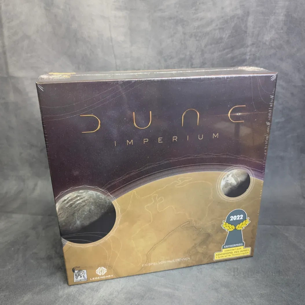 Das Bild zeigt eine versiegelte Schachtel von Dune: Imperium von Asmodee, mit stilisierten Dünen, einem Planeten und einem Aufkleber mit dem Preis für das beste Strategiespiel 2022 unten rechts, der die Deckbuilding-Mechanik hervorhebt.