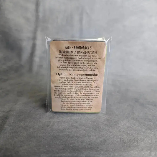 Eine Packung mit der Aufschrift Fate: Die Verteidiger von Grimheim + Helden Erweiterung Sindra & Finkel + Promo Packs von Elznier Games steht aufrecht, in durchsichtigem Plastik verpackt, auf einer grauen, strukturierten Oberfläche mit neutralem grauem Hintergrund.