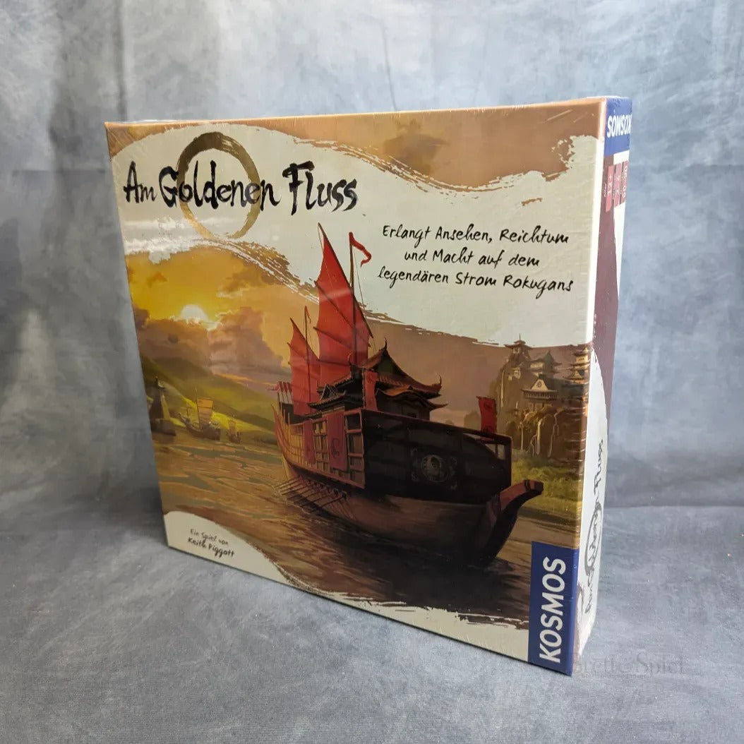 Die Schachtel des Brettspiels Am Goldenen Fluss von Kosmos zeigt ein traditionelles asiatisches Flussschiff auf einem Fluss mit goldenem Sonnenuntergang, während die Spieler zwischen Bergen und Dörfern im Hintergrund ein Handelsimperium aufbauen.