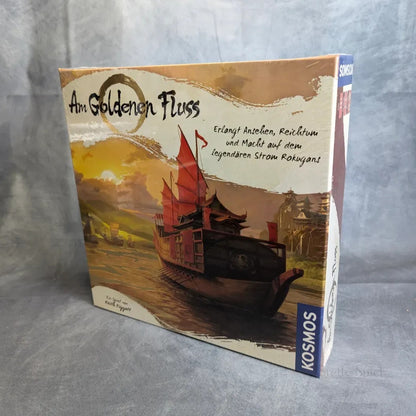 Die Schachtel des Brettspiels Am Goldenen Fluss von Kosmos zeigt ein traditionelles asiatisches Flussschiff auf einem Fluss mit goldenem Sonnenuntergang, während die Spieler zwischen Bergen und Dörfern im Hintergrund ein Handelsimperium aufbauen.