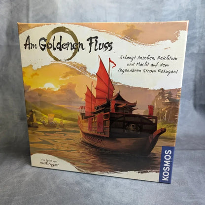 Das Brettspiel Am Goldenen Fluss von Kosmos zeigt eine Schachtel mit einem großen hölzernen Schiff mit roten Segeln, das bei Sonnenuntergang auf einem goldenen Fluss segelt, hinter dem sich Berge erheben - ein Hinweis auf die epische Aufgabe, Ihr Handelsimperium aufzubauen.