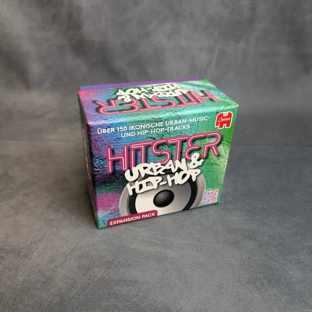 Eine bunte Schachtel mit der Aufschrift Hitster - Urban & Hip-Hop von Jumbo Spiele steht auf einer grauen Fläche. Die Verpackung des Partyspiels ist mit einer Lautsprechergrafik und einem deutschen Text versehen, der über 150 kultige Urban- und Hip-Hop-Tracks hervorhebt.