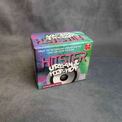 Eine bunte Schachtel mit der Aufschrift Hitster - Urban & Hip-Hop von Jumbo Spiele steht auf einer grauen Fläche. Die Verpackung des Partyspiels ist mit einer Lautsprechergrafik und einem deutschen Text versehen, der über 150 kultige Urban- und Hip-Hop-Tracks hervorhebt.