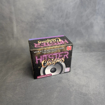 Eine schwarze Box mit der Aufschrift Hitster: Christmas von Jumbo Spiele mit dem Neon-Text "Weihnachten" und "Expansion Pack" steht auf einer grauen Fläche und hebt eine Musik-Challenge mit über 150 Weihnachtsliedern und einer Lautsprechergrafik hervor.