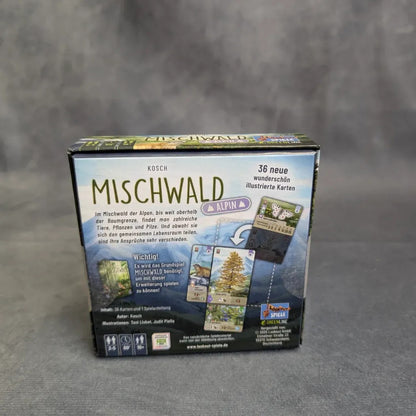 Eine Schachtel von Asmodee's Mischwald - Alpin steht vor einem grauen Hintergrund, der Illustrationen der Alpenflora und -fauna zeigt. Der deutsche Text beschreibt das Spiel, das 36 neue illustrierte Karten mit Bäumen, Tieren und Pflanzen enthält.