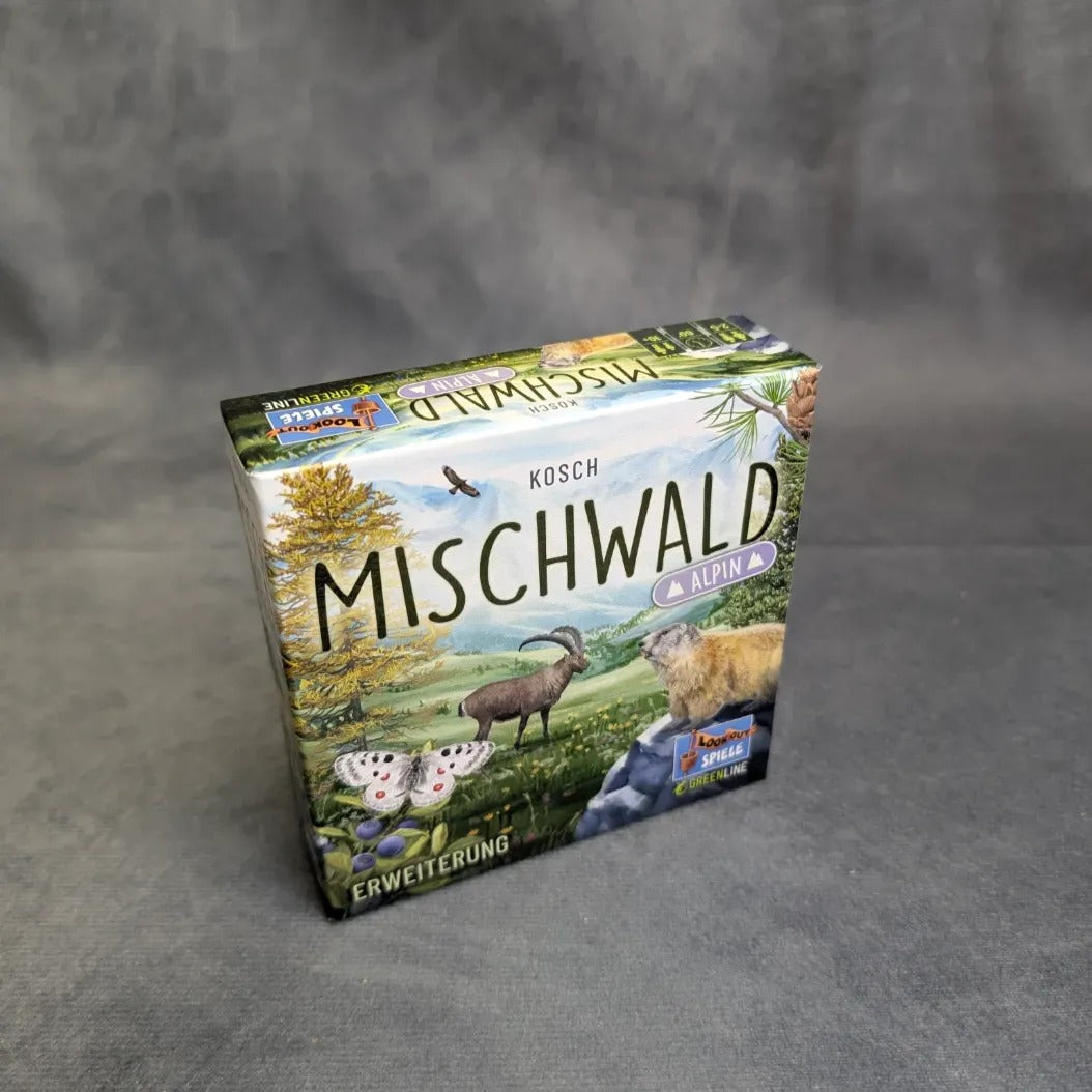 Eine Schachtel Mischwald - Alpin von Asmodee steht auf einer grauen Fläche mit neutralem Hintergrund und zeigt auf dem Deckel illustrierte Alpentiere, Bäume und Berge.