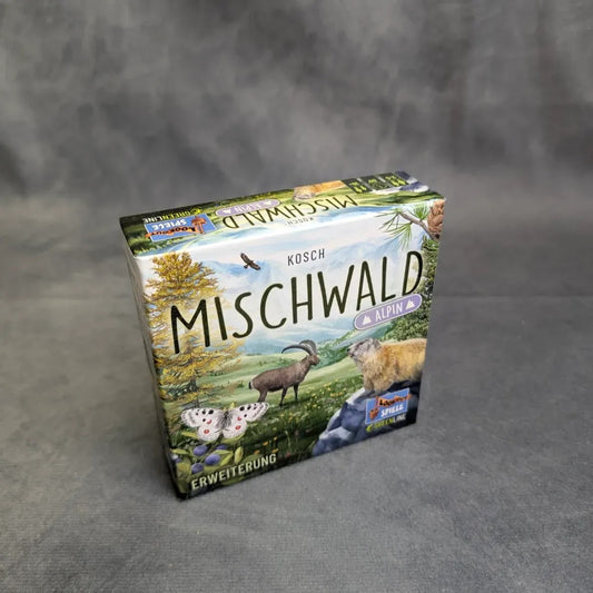 Eine Schachtel Mischwald - Alpin von Asmodee steht auf einer grauen Fläche mit neutralem Hintergrund und zeigt auf dem Deckel illustrierte Alpentiere, Bäume und Berge.