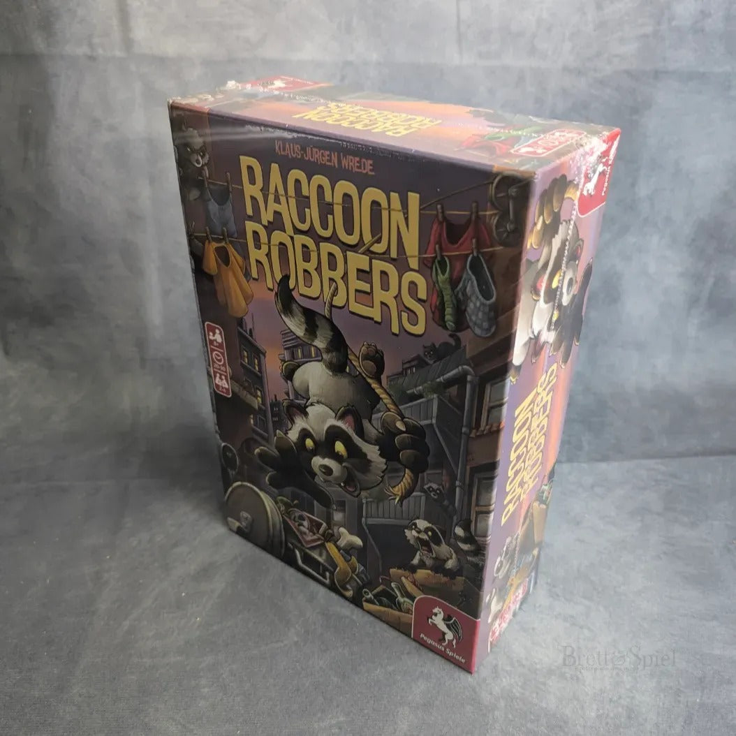 Eine eingeschweißte Pegasus Spiele Brettspiel-Box, Raccoon Robbers, zeigt Cartoon-Waschbären, die durch eine nächtliche Stadt schleichen, mit Taschen voller Beute, Gebäuden und Wäscheleinen im Hintergrund.