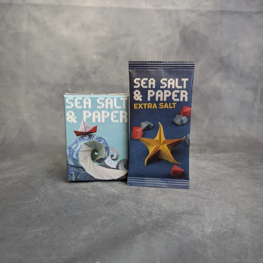 Zwei Spielschachteln Sea,Salt & Paper + Extra Salt Erweiterung von MM-Spiele sind auf grauem Hintergrund abgebildet; die linke zeigt eine Origami-Welle, die rechte, mit Extra Salt beschriftete, einen Origami-Stern und kleine rote Papierformen.