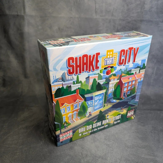 Die Schachtel Shake That City von Boardgame Circus zeigt lebendige Stadtgebäude und Straßen, mit dem Titel des Spiels in fetten roten und gelben Buchstaben vor einem grauen Hintergrund.