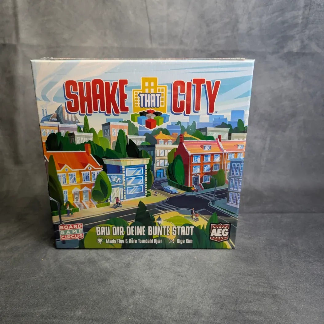 Shake That City von Boardgame Circus ist ein verpacktes Städtebau-Spiel mit lebendigen Gebäude- und Grün-Illustrationen auf dem Cover, die vor einem grauen, strukturierten Hintergrund dargestellt sind.