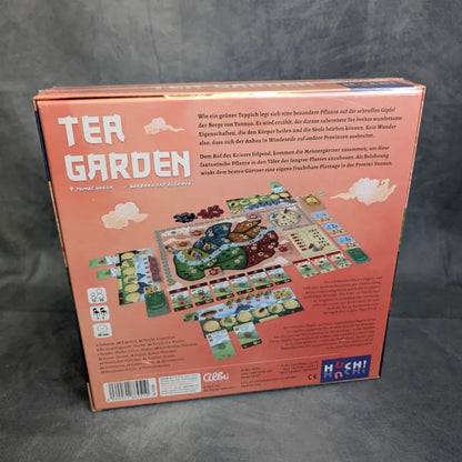 Die Rückseite der Schachtel des Brettspiels Tea Garden aus dem Huch Verlag zeigt einen lebendigen Aufbau mit Karten, Spielsteinen, Plättchen und Spielbrett, dazu deutsche Texte über den Teeanbau und Logos, alles vor einem rosa Hintergrund.