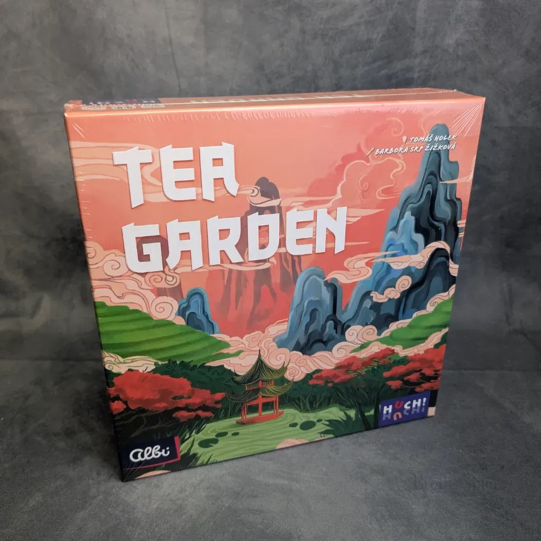 Die Schachtel des Brettspiels Tea Garden vom Huch Verlag zeigt lebendige Bilder von Bergen, Wolken und einer Pagode inmitten einer üppigen grünen Landschaft - die perfekte Kulisse für dieses Strategiespiel über den Teeanbau. Die Verlagslogos befinden sich in den unteren Ecken.