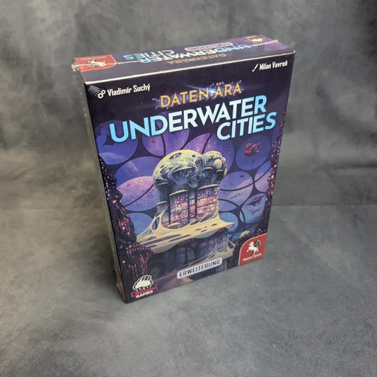 Eine Schachtel von Underwater Cities: Daten-Ära (Erweiterung) von Pegasus Spiele ruht auf einer grauen Fläche, auf deren Vorderseite bunte Unterwasser-Grafiken und mehrere Logos abgebildet sind.
