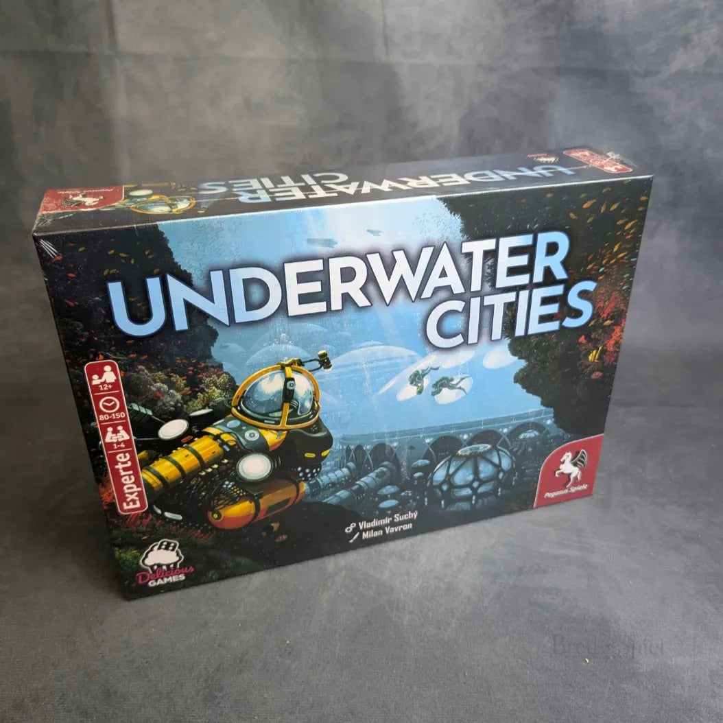 Eine Schachtel mit den Unterwasserstädten von Pegasus Spiele, die ein gelbes U-Boot und Unterwasserkuppeln inmitten eines leuchtend blauen Ozeans mit Korallen und Meeresbewohnern zeigen - ideal für Städtebaufans.