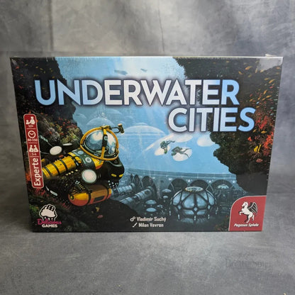 Die Schachtel von Underwater Cities zeigt eine futuristische Unterwasserszene mit einem gelben U-Boot, Blasen, markanten Stadtstrukturen und dem fetten blauen Titel. Das Logo von Pegasus Spiele erscheint auch auf dem Cover dieses Strategiespiels.