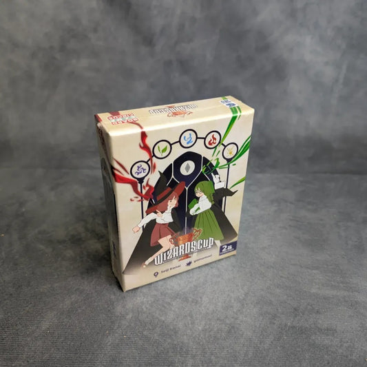Wizards Cup von Boardgame Circus ist ein verpacktes Kartenspiel auf einer grauen Oberfläche. Die Verpackung zeigt zwei animierte Charaktere in strategischen Duellen, die Zaubersprüche wirken, mit lebendigen magischen Symbolen über ihnen.