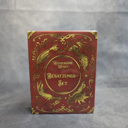 Eine rote Schachtel mit goldenen Motiven von magischen Kreaturen wie einem Drachen und einer Seeschlange. Der deutsche Text lautet "Wundersame Wesen + Besatzungs-Set + Gigantische Bestien Erweiterung" von Strohmann Games. Grauer Hintergrund.