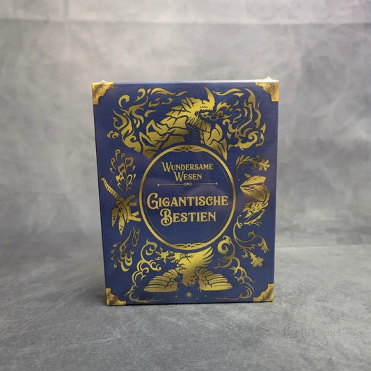 Eine blau-gold verzierte Schachtel mit Abbildungen fantastischer Kreaturen, betitelt "Wundersame Wesen + Besatzungs-Set + Gigantische Bestien Erweiterung" von Strohmann Games, steht aufrecht vor einem grauen Hintergrund.