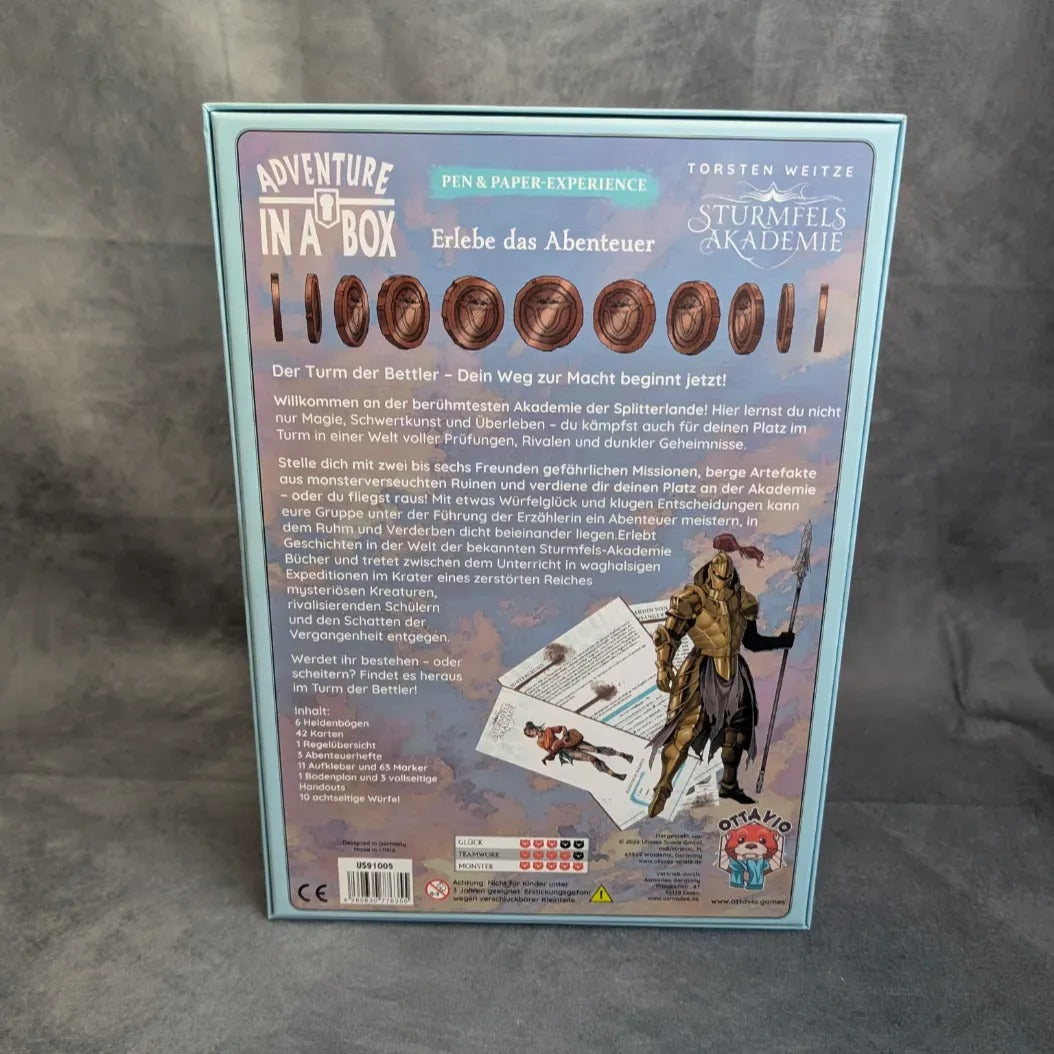 Die Rückseite der "Adventure in a Box: Sturmfels Akademie - Der Turm der Bettler" der Ulisses Spiele GmbH ist mit deutschem Text, einem Pen & Paper Experience-Banner, Ritter- und Akademie-Artwork aus der Splitterlande und Logos auf blau-grünem Hintergrund versehen.
