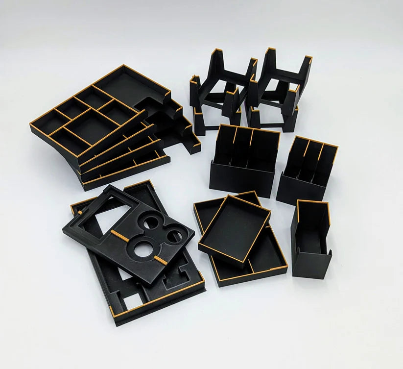 Mehrere schwarze rechteckige und quadratische Tabletts, Boxen und Organizer mit orangefarbenen Rändern aus dem 3D-Inset Dune Imperium Insert sind auf einer weißen Fläche angeordnet. Diese Elemente haben Fächer und Schlitze für die organisierte Aufbewahrung von Spielen.