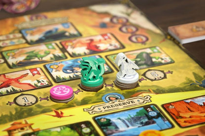 Eine Nahaufnahme des Brettspiels Glutherz von Mindclash Games zeigt Drachen- und Geisterfiguren auf Plättchenstapeln, ein rosafarbenes Pfotenabdruckplättchen, alles auf einem PRESERVE-Feld, mit Karten und illustrierten Abschnitten im Hintergrund.