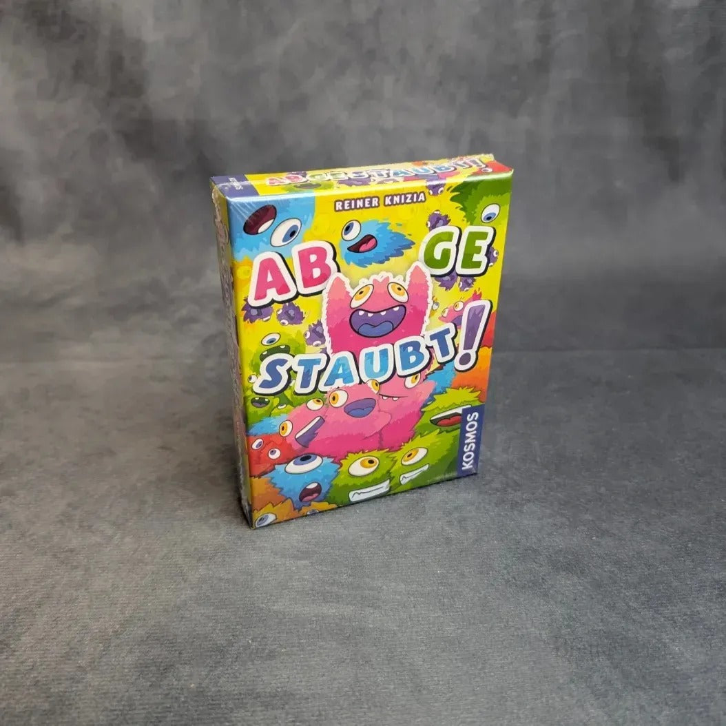 Das Kartenspiel „Abgestaubt“ von Kosmos wird in einer farbenfrohen Schachtel mit verspielten Monstermotiven in Grün, Rosa und Gelb auf grauem Hintergrund geliefert – eine tolle Wahl für dein nächstes Kartenspiel oder deine nächste Staubfussel-Jagd.