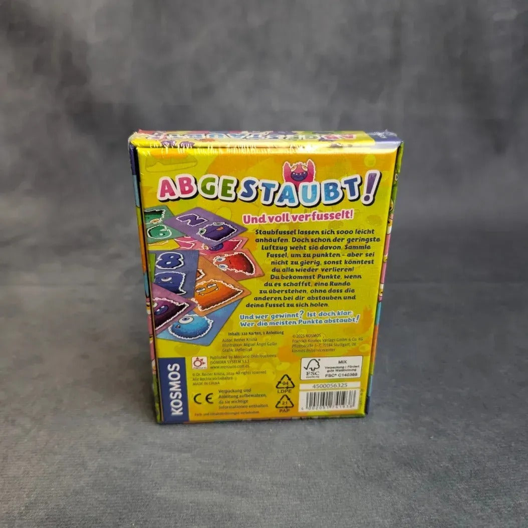 Die „Kosmos Abgestaubt“-Schachtel steht mit der Rückseite zur Kamera und zeigt deutschen Text, farbenfrohe Grafiken sowie Abbildungen von nummerierten, staubbedeckten Spielsteinen für eine spielerische Staubfussel-Jagd.